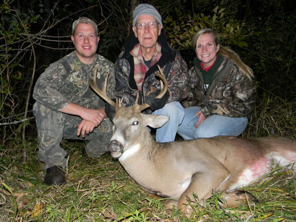 Three people in camouflage and outdoor clothing kneel behind a large deer with antlers, showcasing a successful deer hunting trip in a wooded area—perfect for those using Michigan Hunting Blinds. - Michigan Hunting Blinds