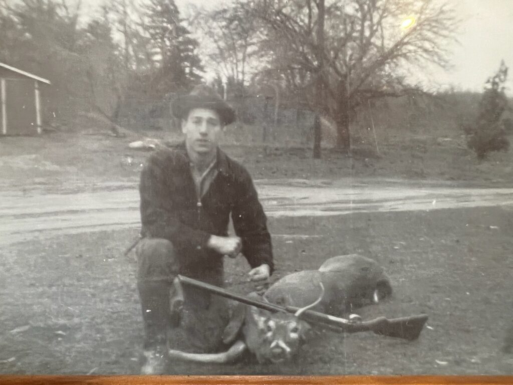 A man wearing a hat kneels outdoors beside a rifle and a deer on the ground, with trees, a small building, and Michigan Hunting Blinds visible in the background. - Michigan Hunting Blinds