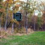 The Big Country Platinum 360° 6X7 hunting blind from Redneck Deer Blinds stands on metal stilts among trees, surrounded by grass and autumn foliage in a peaceful forested area. - Michigan Hunting Blinds