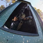 A hunter aims a compound bow out the window of a Big Country Platinum 360° 6X7 camouflaged deer blind, preparing to shoot. - Michigan Hunting Blinds