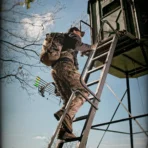 A person in camouflage climbs a metal ladder to the Big Country Platinum 360° 6X7 deer hunting blind, carrying a backpack and a bow with arrows. - Michigan Hunting Blinds