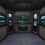 Interior of the Big Country Platinum 360° 6X7 black, soundproofed deer blind with padded walls, four windows overlooking grassy views, and an electronic device mounted below one window. - Michigan Hunting Blinds